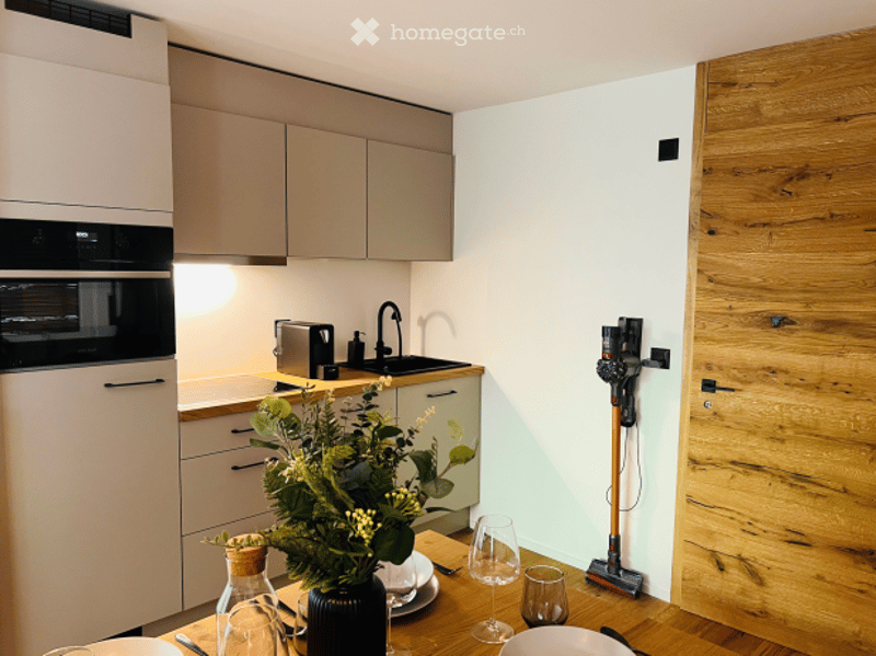 Modern kitchen with light-colored cabinets, a built-in oven, a sink with a faucet, and a dining area with a wooden table and chairs. A vase with flowers and wine glasses are on the table, and a vacuum cleaner is mounted on the wall.