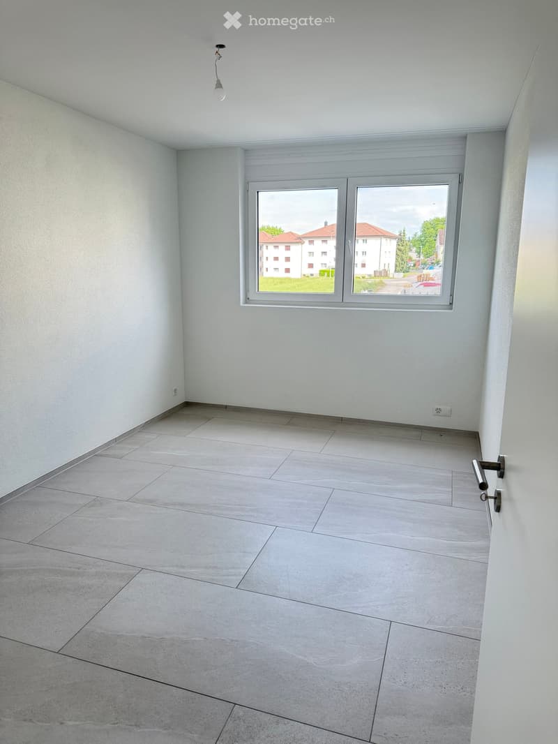 Empty room with light grey tiled floor, white walls, and a large window with a view of buildings and greenery outside.
