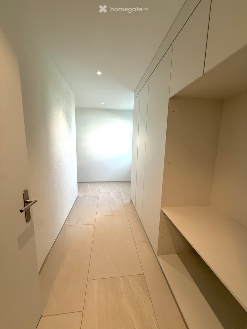 Modern hallway with white walls, light-colored tiled floor, built-in shelving on one side, and recessed ceiling lighting.