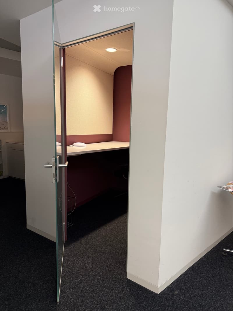 A small, modern office space with a glass door, a white desk, and a chair. The room has a dark carpeted floor and white walls.