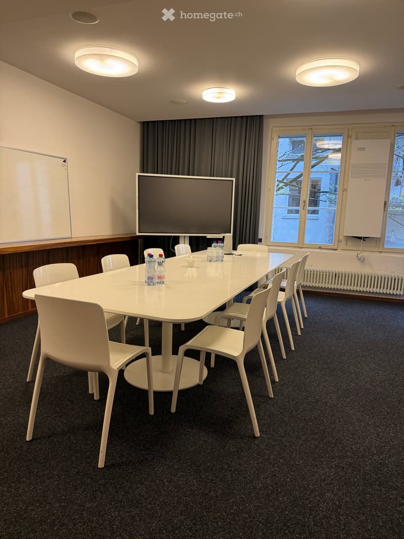 Modern conference room with a large white table, multiple chairs, a large screen, and several water bottles on the table. The room has large windows with curtains and is well-lit with ceiling lights.