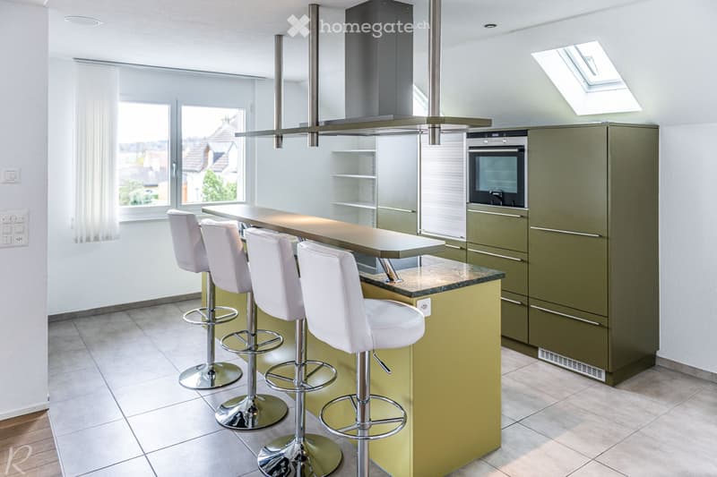 Modern kitchen with a bar counter, four white stools, green cabinets, and an integrated oven. The space features a skylight and large windows offering a view of the neighborhood.