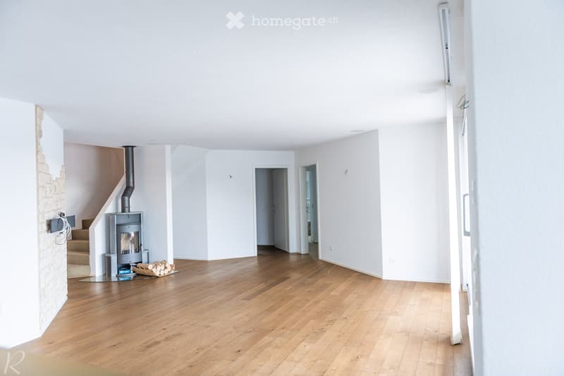 Empty room with wooden flooring, white walls, and a staircase leading upwards. A wood-burning stove is present, along with a small basket of firewood.