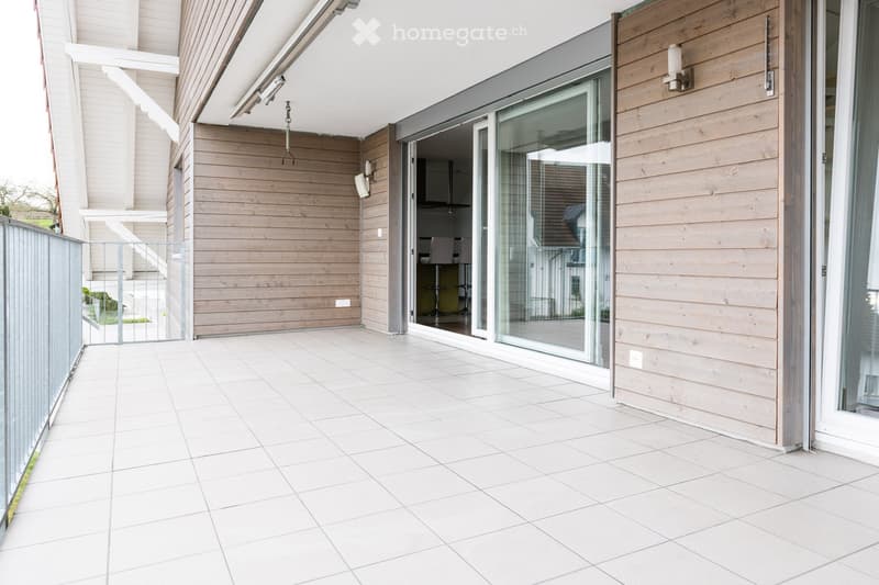 Modern balcony with white tiled flooring, large glass sliding doors leading to the interior, wooden panel walls, and a metal gate.