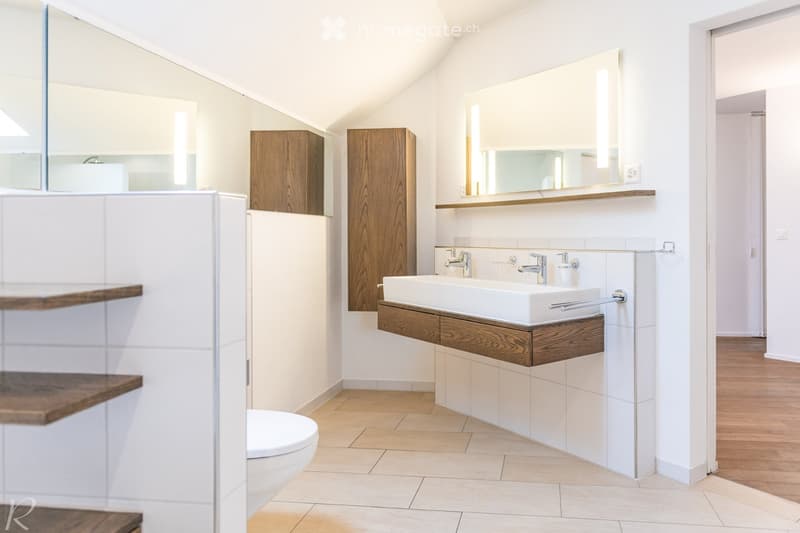 Modern bathroom with white tiles, wooden cabinets, a large sink with chrome faucets, and a large mirror above it. The floor is tiled, and there is a toilet visible on the left side.