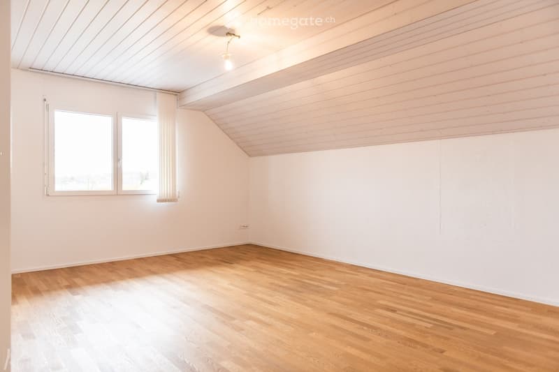 Empty room with wooden flooring, white walls, and a slanted ceiling. Two windows with white frames and curtains allow natural light to enter.