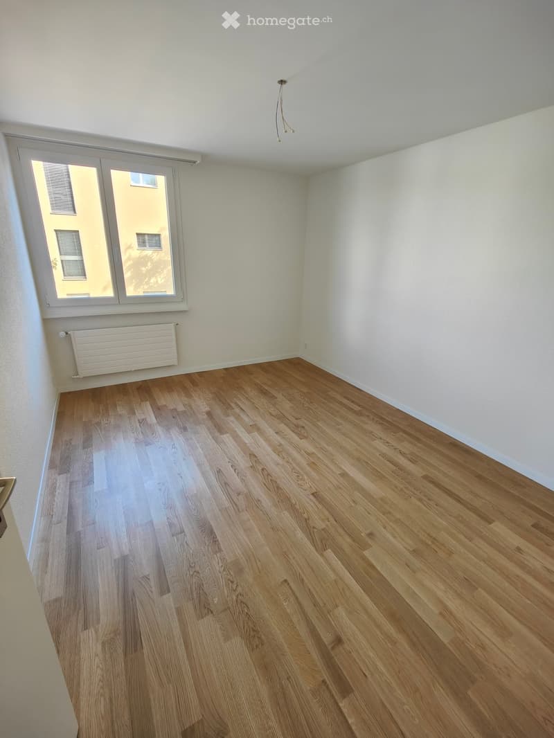 Empty room with wooden flooring, white walls, and a large window with blinds. A radiator is visible below the window.