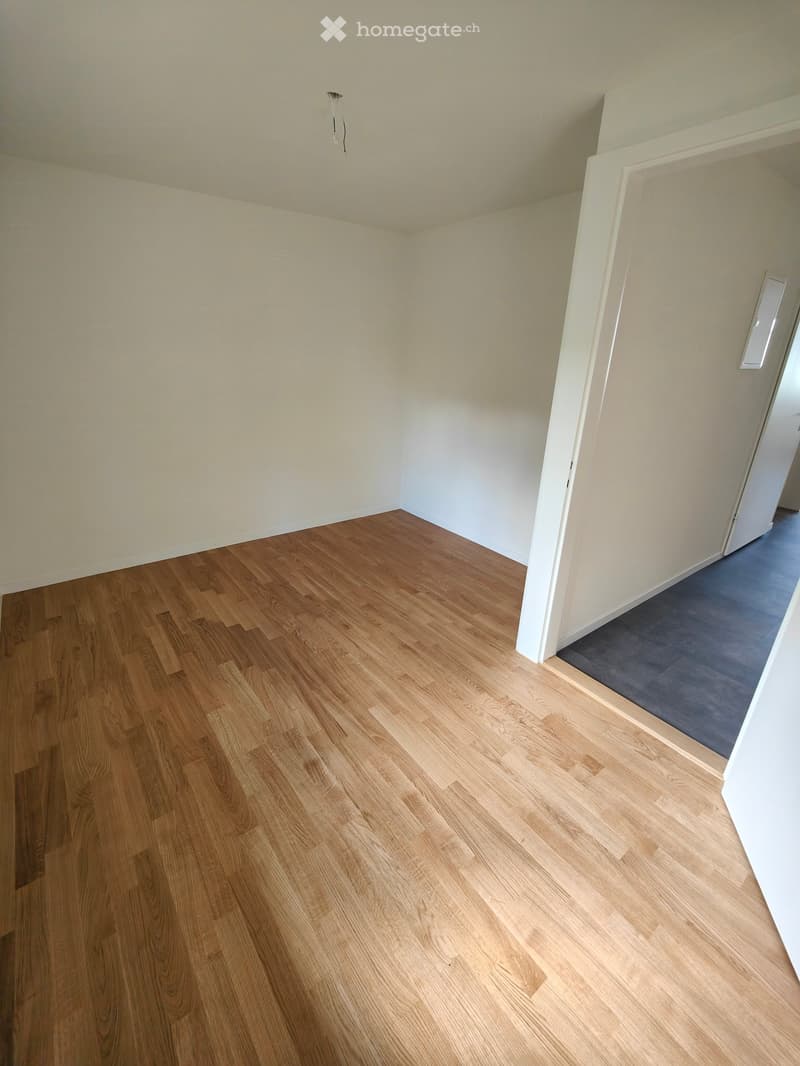 Empty room with light wooden flooring and white walls, featuring a doorway leading to another room with a gray carpet.