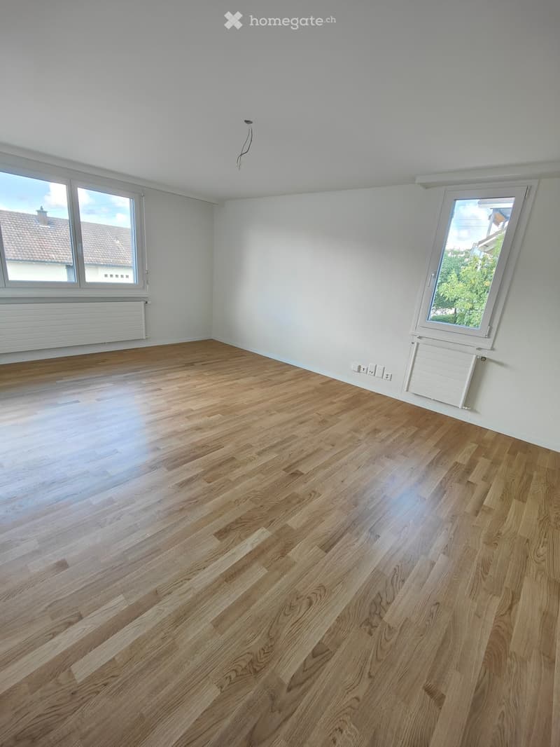 Empty room with light wooden flooring, white walls, and large windows offering a view of the outside. Radiators are visible under the windows.