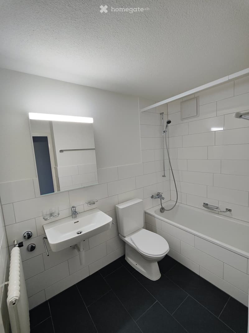 Modern bathroom with white tiles, a sink, toilet, bathtub, and a large mirror with built-in lighting. Features a dark tiled floor and a towel on the wall.