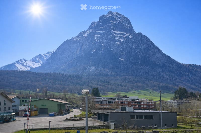 A small town or village with several buildings, including a garage and a multi-story structure, set against a backdrop of snow-capped mountains and a clear blue sky.