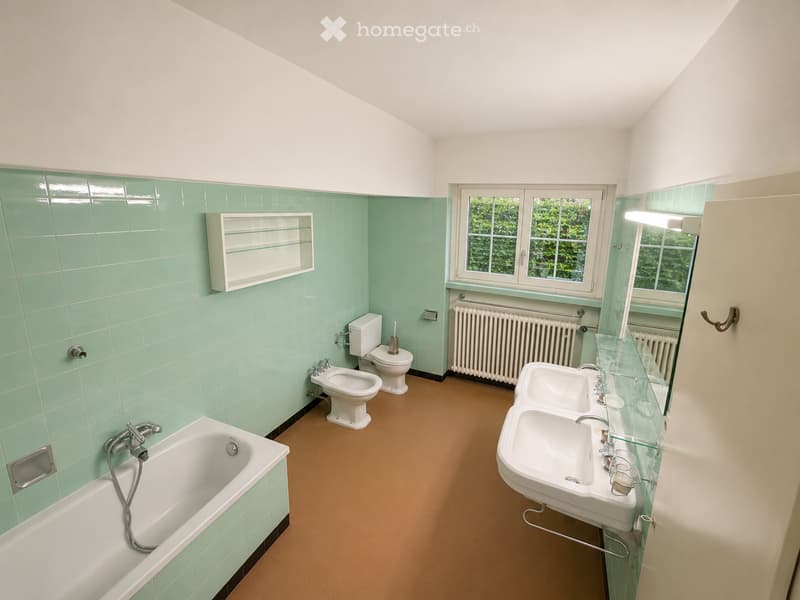 Bathroom with mint green tiles, white fixtures, bathtub, double sink, toilet, bidet, radiator, and a window with a view of greenery.