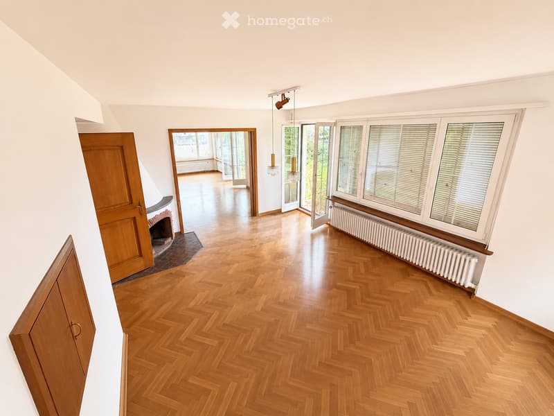 Bright hallway with wooden herringbone flooring, white walls, large windows, and a visible fireplace area.