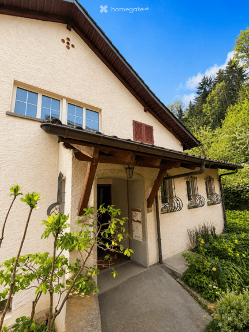 A charming, light-colored house with a wooden entrance canopy, multiple windows, and a small garden area with greenery and flowers.