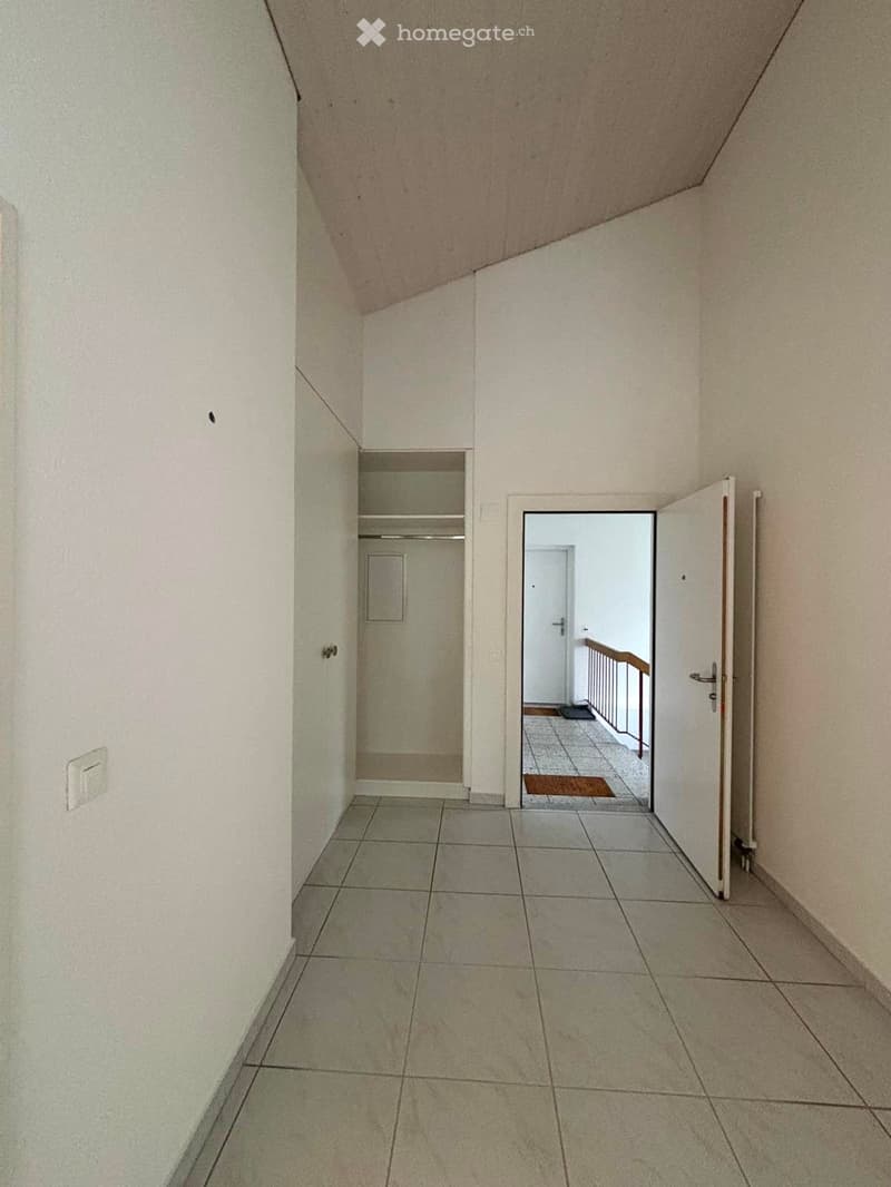 Empty hallway with tiled floor, white walls, and an open door leading to another room with a visible staircase and railing.