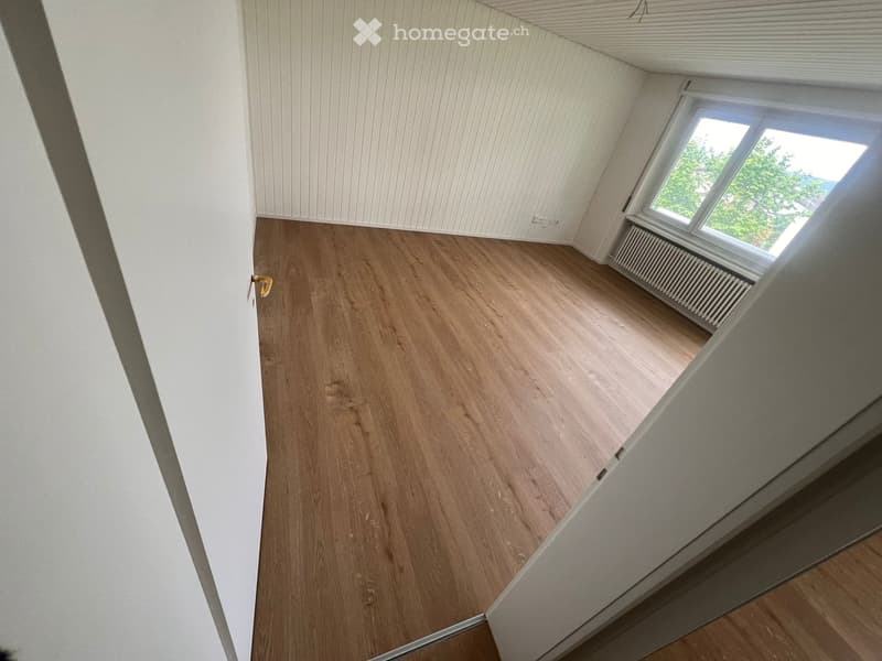 Empty attic room with wooden flooring, white walls, and a single window showing greenery outside.