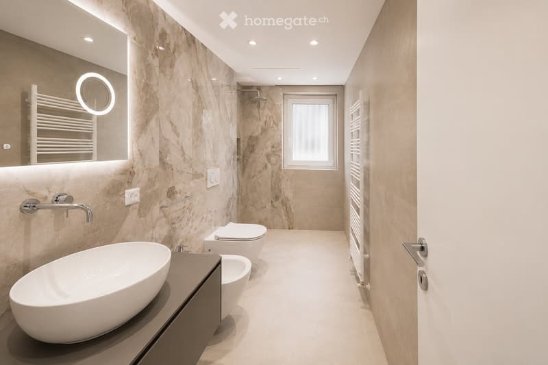 Modern bathroom with marble walls, a large oval sink, a toilet, a bathtub, a shower area, and a window. Features include a built-in radiator, a round mirror with LED lighting, and sleek chrome fixtures.