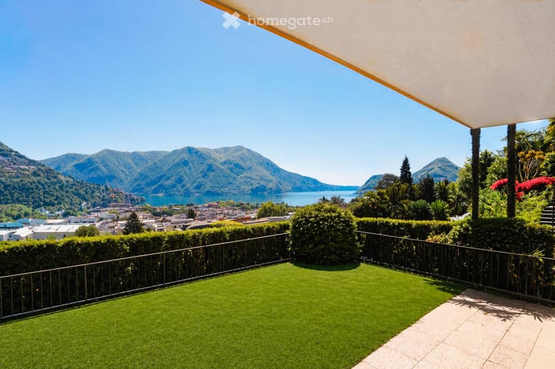 A spacious balcony with artificial grass, a metal railing, and a panoramic view of a lake and surrounding mountains.