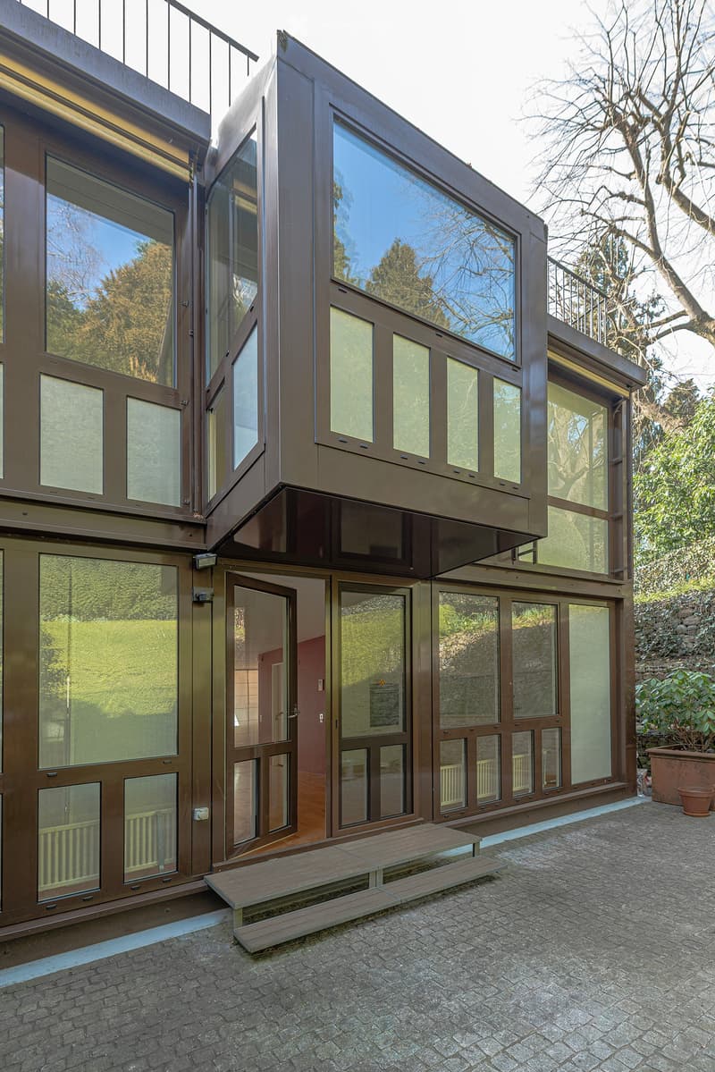 Modern building with large glass windows and brown exterior. The entrance is open, revealing a wooden step leading up to the entrance.