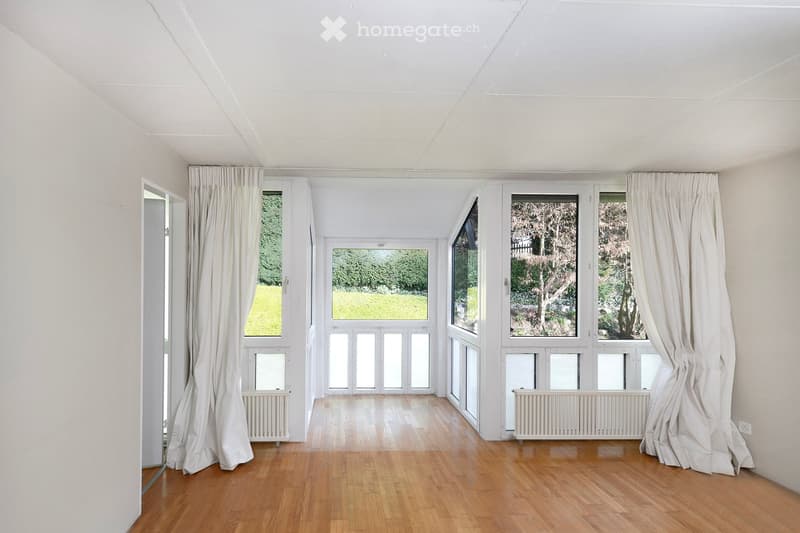 An empty room with white walls, wooden floor, and large windows with white curtains. There are white radiators under the windows.