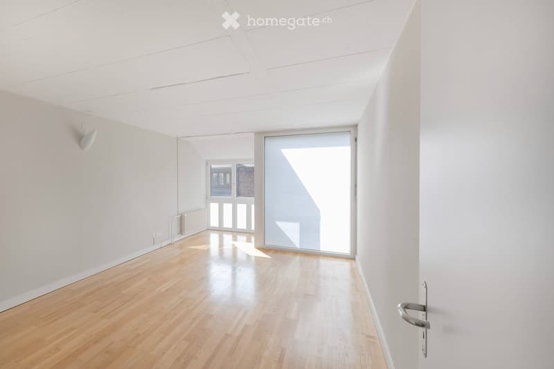 Empty room with light wooden flooring, white walls, and large windows allowing natural light. The space is unfurnished and features a simple, modern design.