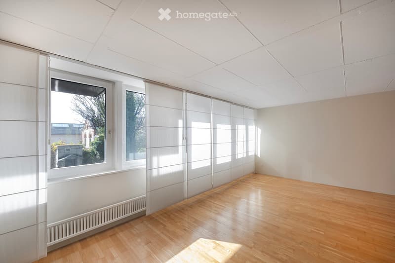 An empty room with white walls, wooden floor, and large windows with white blinds. There is a white radiator on the wall.