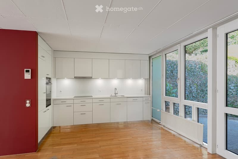 Modern kitchen with white cabinets, red accent wall, wooden floor, and large windows.