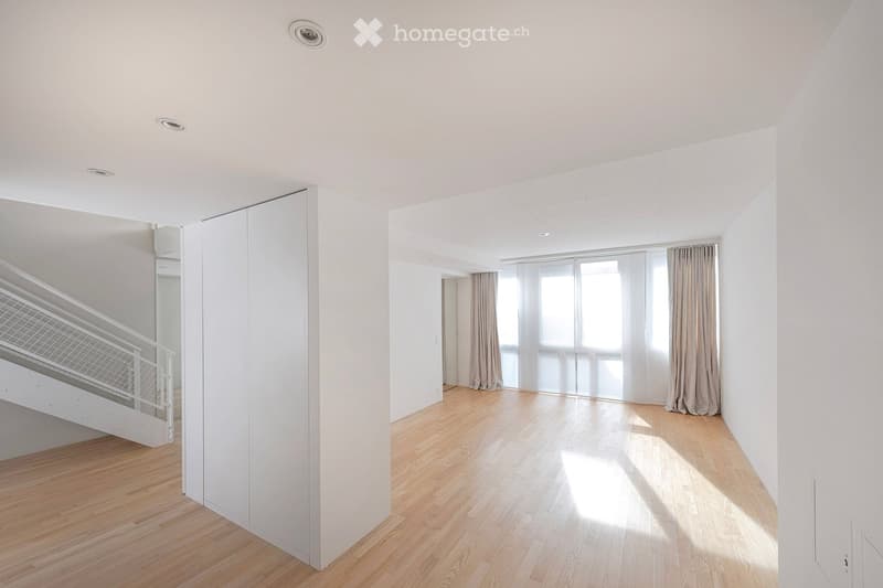 Bright, empty room with wooden flooring, white walls, and large windows with curtains. A staircase leads to an upper level.