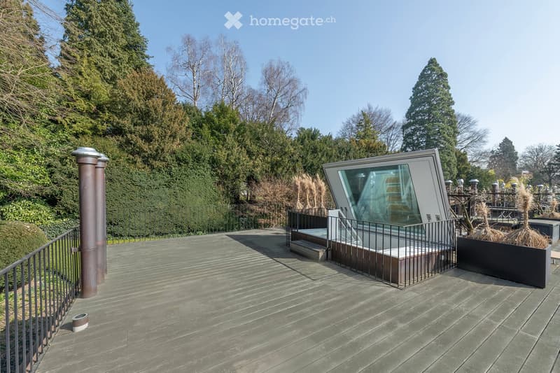 Rooftop terrace with wooden deck, black railing, and a tub. View of the city and mountains.