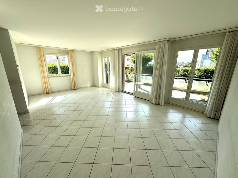Empty room with white tiled floor, large windows with curtains, and sliding glass doors leading to a balcony.