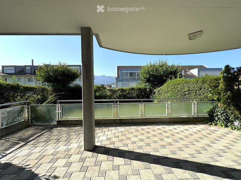 Balcony with tiled floor, metal railing, and glass panels, overlooking a residential area with trees and mountains in the background.