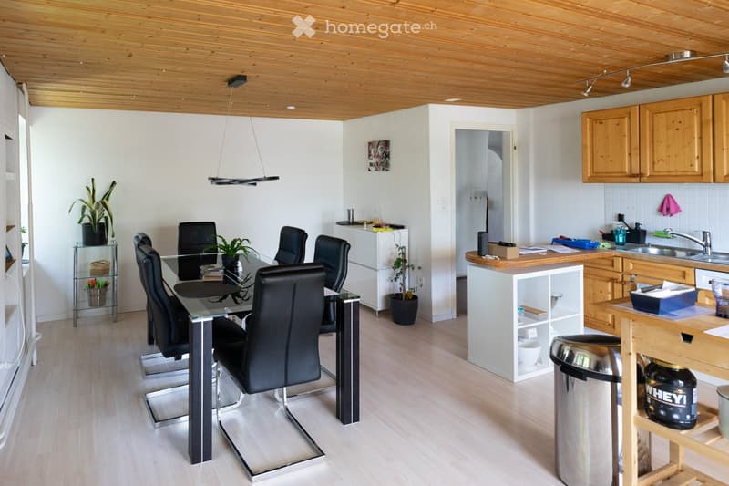 Modern open-plan kitchen and dining area with light wooden flooring, white walls, and a wooden ceiling. Features a glass dining table with black chairs, a wooden kitchen island, and various kitchen cabinets and appliances.