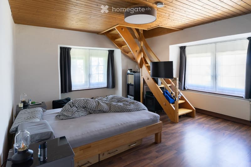 Cozy bedroom with wooden floors, slanted wooden ceiling, large windows, bed with grey bedding, nightstand with lamp, mirror, TV, staircase leading to loft area, and dreamcatcher decoration.