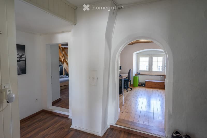 A bright, open-plan room with wooden flooring, white walls, and arched doorways leading to other spaces. Visible objects include a desk with a chair, a small stool, a radiator, and a picture frame on the wall.