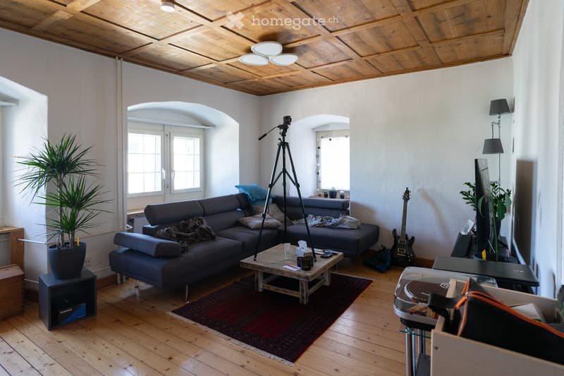 Living room with wooden floor, exposed wooden ceiling, large windows, grey sectional sofa, coffee table, guitar, tripod, laptop, and plants.