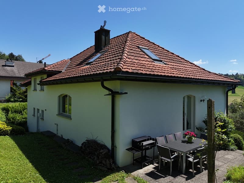 A small, modern house with a red-tiled roof and white walls, featuring a patio area with a dining table, chairs, and a grill. The surrounding area includes a well-maintained lawn and garden.