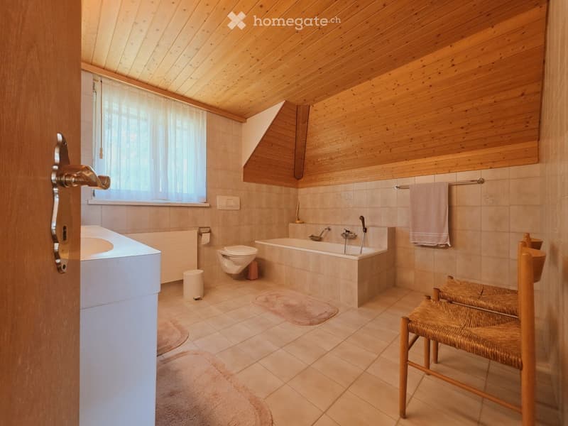 Bathroom with wooden ceiling and walls, tiled floor, bathtub, toilet, sink, chair, towel, and window with curtain.