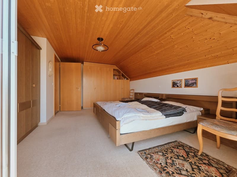 Cozy attic bedroom with wooden slanted ceilings, a large bed with white bedding, a wooden chair, a wardrobe, and a patterned rug.