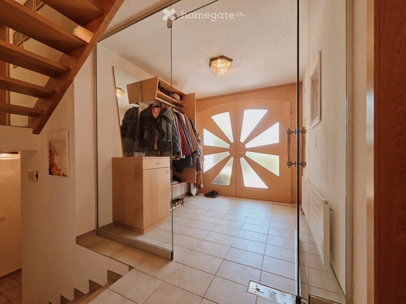 Modern hallway with tiled floor, wooden staircase, built-in closet with hanging coats, and a large glass door with a sun-ray design.