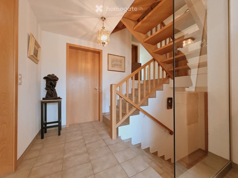 Spacious hallway with wooden staircase, glass railing, pendant light, small table with bust, framed artwork, and tiled floor.