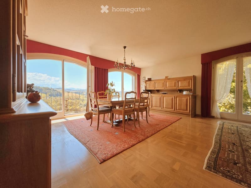 Elegant dining room with wooden flooring, large windows offering scenic views, a dining table surrounded by chairs, a wooden cabinet, and a chandelier hanging from the ceiling.