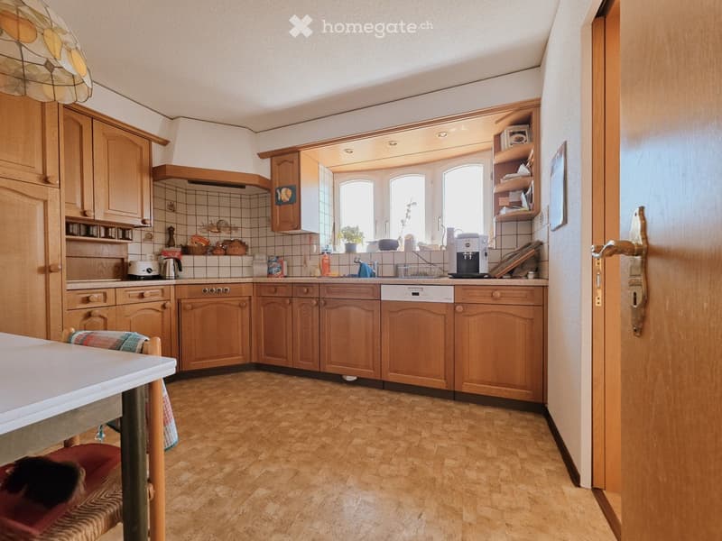 The kitchen features wooden cabinets, a tiled backsplash, and a wooden floor. Visible appliances include an oven, dishwasher, coffee maker, and a sink with a faucet. There is a small table with a chair, and shelves with various items.