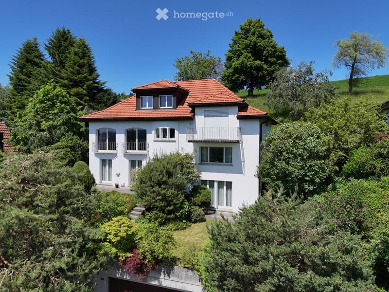 A two-story white house with a red-tiled roof, surrounded by lush greenery and trees. Features include multiple windows, balconies, and a visible garage entrance.