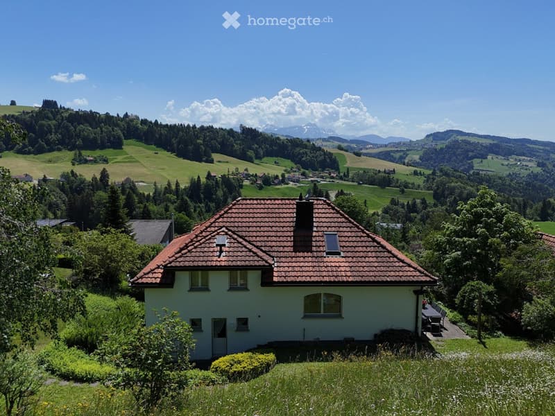 A single-story house with a red-tiled roof and light yellow walls, surrounded by lush greenery and a scenic mountain backdrop.