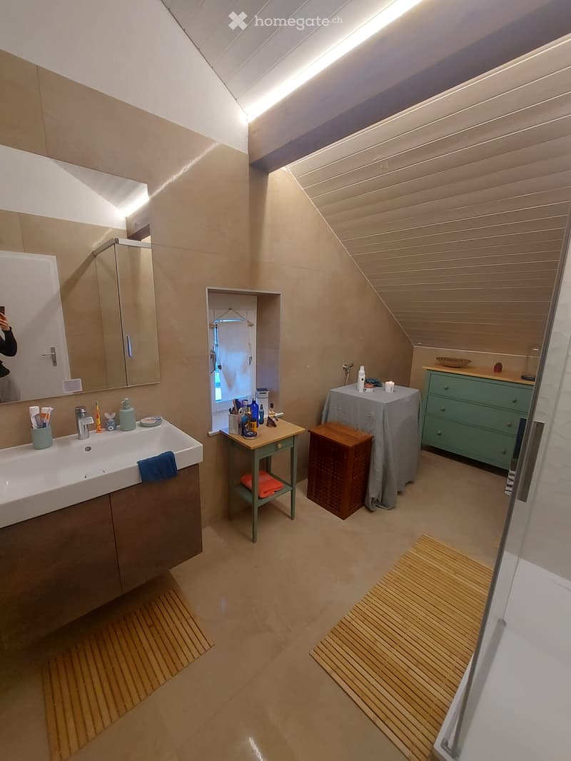 Modern bathroom with a large sink, mirror, and wooden floor. Features include a small table with toiletries, a stool, a covered table, and a green cabinet.