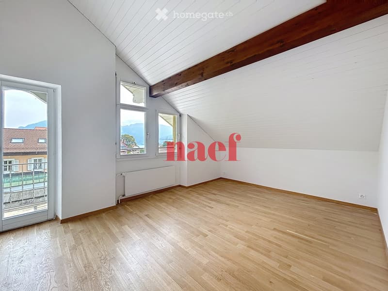 Empty room with wooden flooring, white walls, and large windows offering a scenic view. The room features exposed wooden beams on the ceiling and a radiator along one wall.