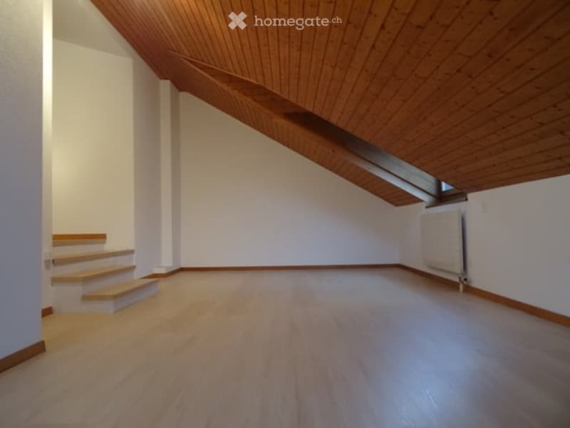 Empty room with wooden flooring, white walls, and a wooden staircase leading up, featuring a slanted wooden ceiling and a small skylight window.