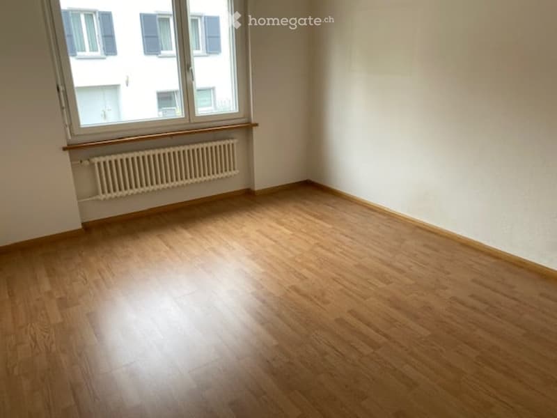 Empty room with wooden flooring, white walls, and a window with a radiator below it.