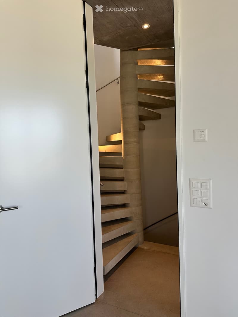 Spiral staircase with concrete steps and a central column, illuminated by recessed lighting, adjacent to a white wall with light switches and a door handle.