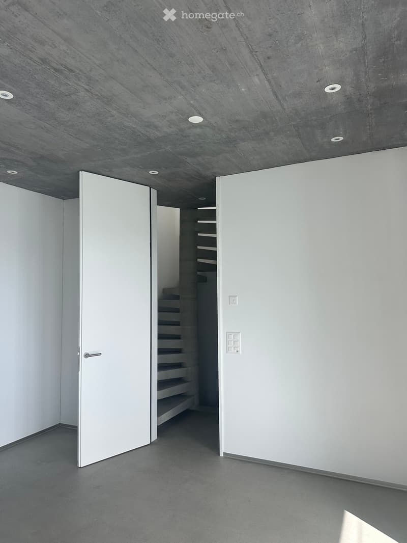 Modern, minimalist hallway with concrete ceiling and floor, white walls, and an open staircase leading upwards.